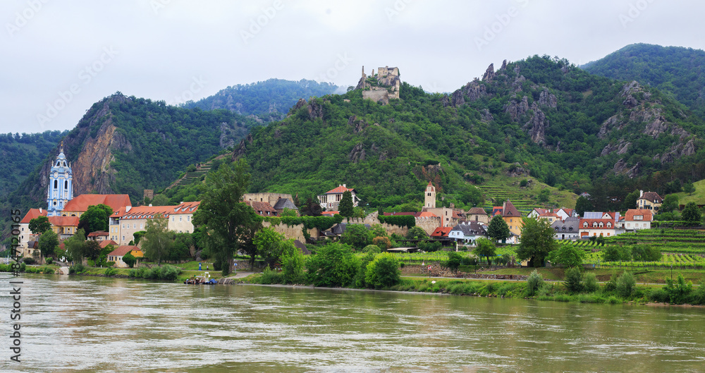 Fototapeta premium Panoramic Scenic view while cruising along the River Danube in Austria. Pretty waterfront village with blue church and quaint cottages, all nestled amongst lush green mountains.