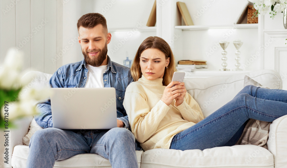 A couple sits on a white sofa in a bright living room, engaged with a laptop and smartphone, respectively, sharing a relaxed moment together. A couple sits on a white sofa in a bright living room, engaged with a laptop and smartphone, respectively, sharing a relaxed moment together.
