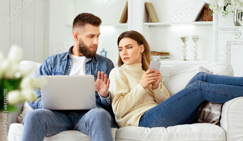 Woman Spying Husband's Laptop, Sitting Together On Sofa With Devices