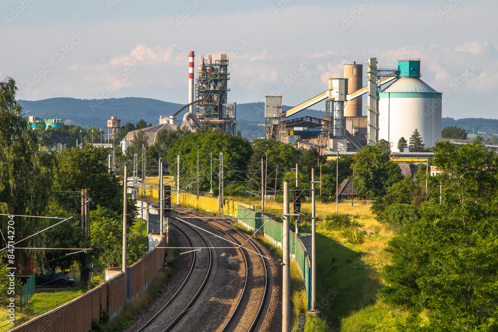 Fototapeta premium The exterior of a cement plant with a factory chimney and a railroad track in the foreground.