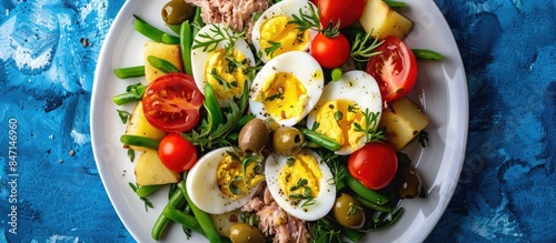 Engaging version: Top view of a white plate showcasing a flavorful Nicoise salad made with canned tuna, tomatoes, boiled eggs, green beans, potatoes, and olives against a blue table background © Lasvu