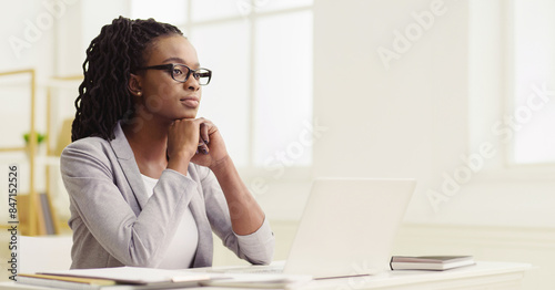 Pensive black woman employee is using her computer in a modern office. The mood is cheerful and productive, with a neat desk and bright space, showing enjoyment in her work.