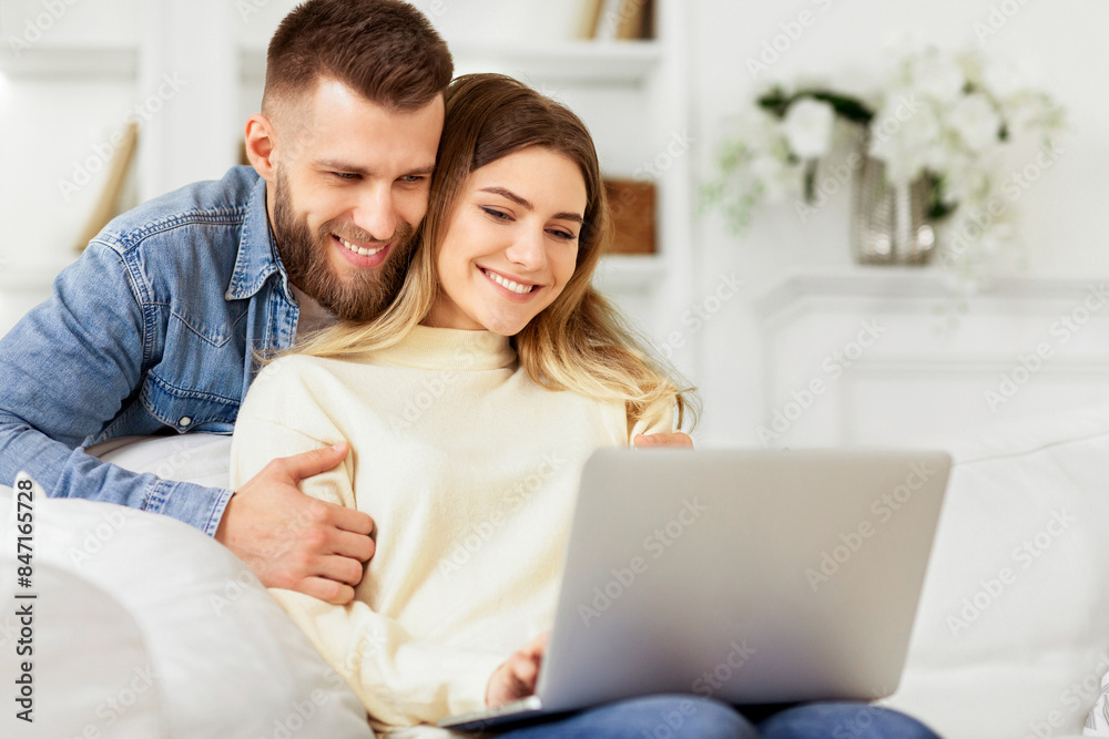 A man and a woman are seated on a couch, focused on a laptop screen in front of them.