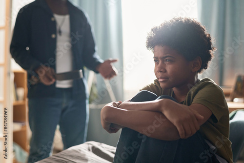 Portrait of African American boy crying and looking away with unrecognizable parent holding belt in background copy space