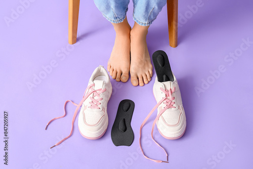 Female legs near orthopedic insoles and sneakers on lilac background, closeup photo