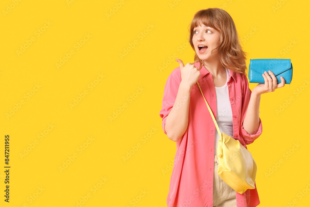 Beautiful young woman with blue wallet pointing at something on yellow background Beautiful young woman with blue wallet pointing at something on yellow background