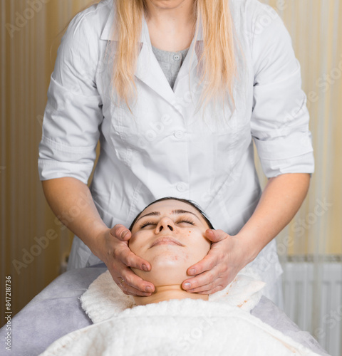 Young woman enjoying facial massage, lying on spa bed indoor at beauty salon.