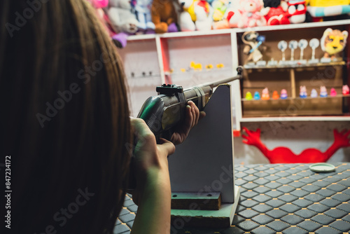 A young girl aiming a rifle at a target in a shooting game booth filled with toy prizes, an over-the-shoulder shot.