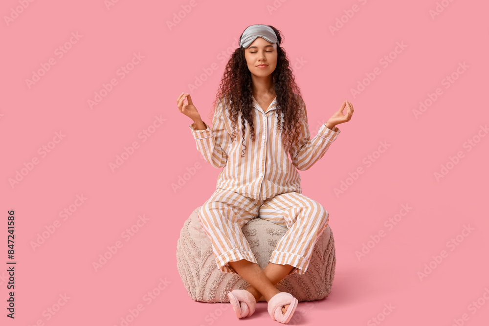 Beautiful young African-American woman in pajamas meditating on pouf against pink background Beautiful young African-American woman in pajamas meditating on pouf against pink background