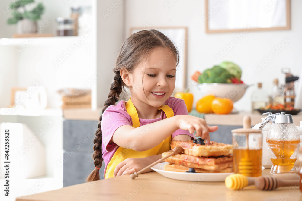 Cute little girl eating waffles with honey in kitchen Cute little girl eating waffles with honey in kitchen