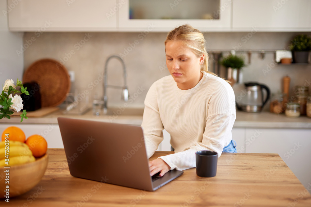 Young woman wearing casual clothes using laptop and drinking coffee in the kitchen