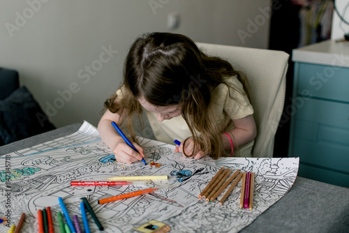 Little girl colors a large coloring book with colored pencils and felt-tip pens at home