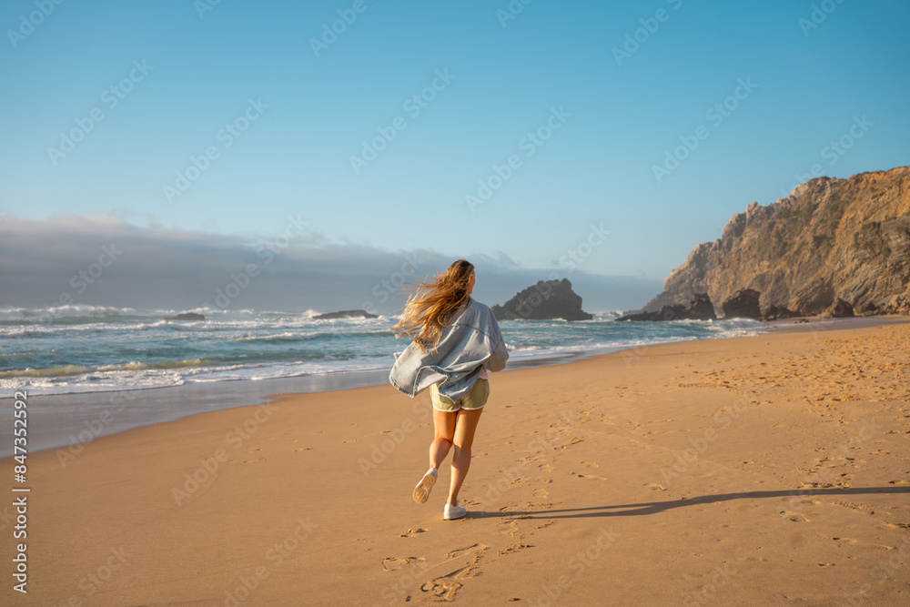 Back view of woman in denim shirt running on beach, hair flying, with ocean view and mountains in the background. Conveys a sense of freedom and summer holidays by the sea. Back view of woman in denim shirt running on beach, hair flying, with ocean view and mountains in the background. Conveys a sense of freedom and summer holidays by the sea.