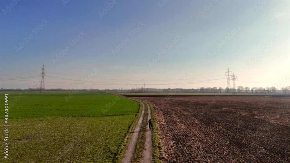 Male cyclist riding gravel bike on country road through farm field in spring view from sky. Man bicyclist cycling on bicycle on rural path across a farmers land in spring, top view. Drone view. Male cyclist riding gravel bike on country road through farm field in spring view from sky. Man bicyclist cycling on bicycle on rural path across a farmers land in spring, top view. Drone view.