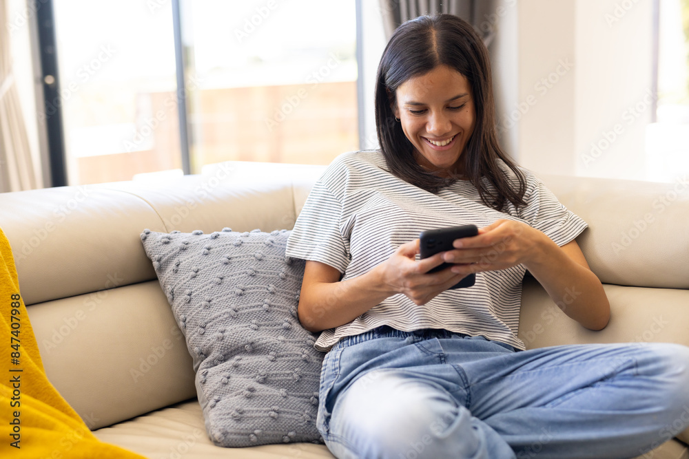 Biracial young woman relaxing on couch, smiling while using smartphone, at home Biracial young woman relaxing on couch, smiling while using smartphone, at home