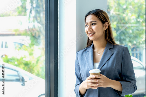 Free Download Young happy Asian businesswoman in a suit holding a cup of delivery coffee, standing confident in the office room background - GFXLoad