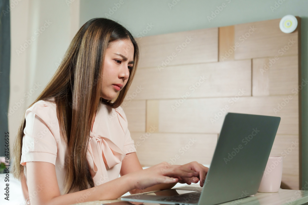 Asian woman with serious expression working on laptop at home. Light-colored blouse, modern home office with wooden wall. Focused and determined, hands typing on keyboard, cup nearby. Asian woman with serious expression working on laptop at home. Light-colored blouse, modern home office with wooden wall. Focused and determined, hands typing on keyboard, cup nearby.