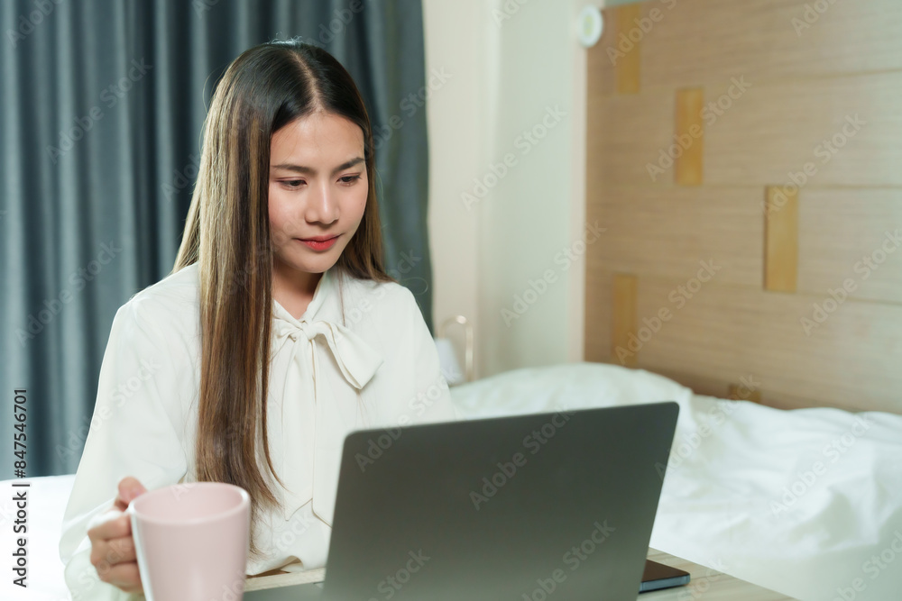 Asian woman in white blouse working on laptop at home, holding pink mug. Indoor home office with curtains and wooden wall. Focused expression, modern workspace, long straight hair, casual setting. Asian woman in white blouse working on laptop at home, holding pink mug. Indoor home office with curtains and wooden wall. Focused expression, modern workspace, long straight hair, casual setting.