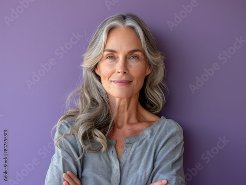A woman with gray hair is smiling and posing for a picture. She is wearing a gray shirt and has her arms crossed