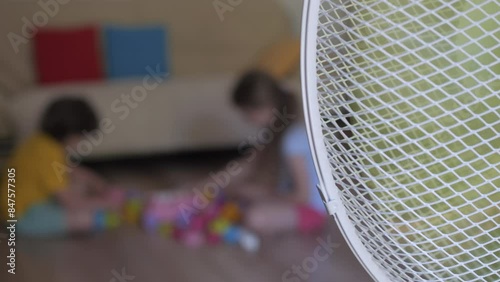 Children Enjoying Cool Wind From Electric Fan at Home at Summer Heat Weather. Children Play Sitting on the floor in Front Fan Hot Day on Home in Living Room. Kids Summer Heat Fresh Cool Blowing Fan.