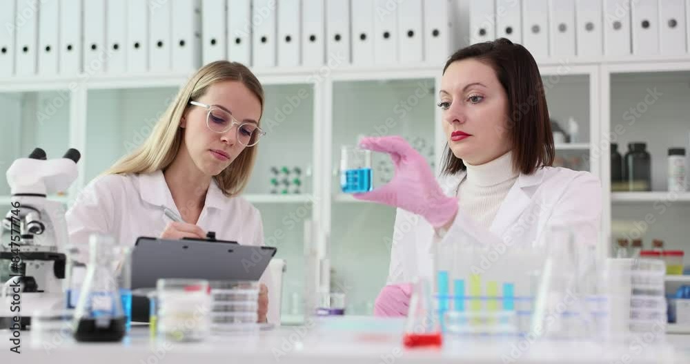 Female scientist describes blue liquid assistant marks notes Female scientist describes blue liquid assistant marks notes