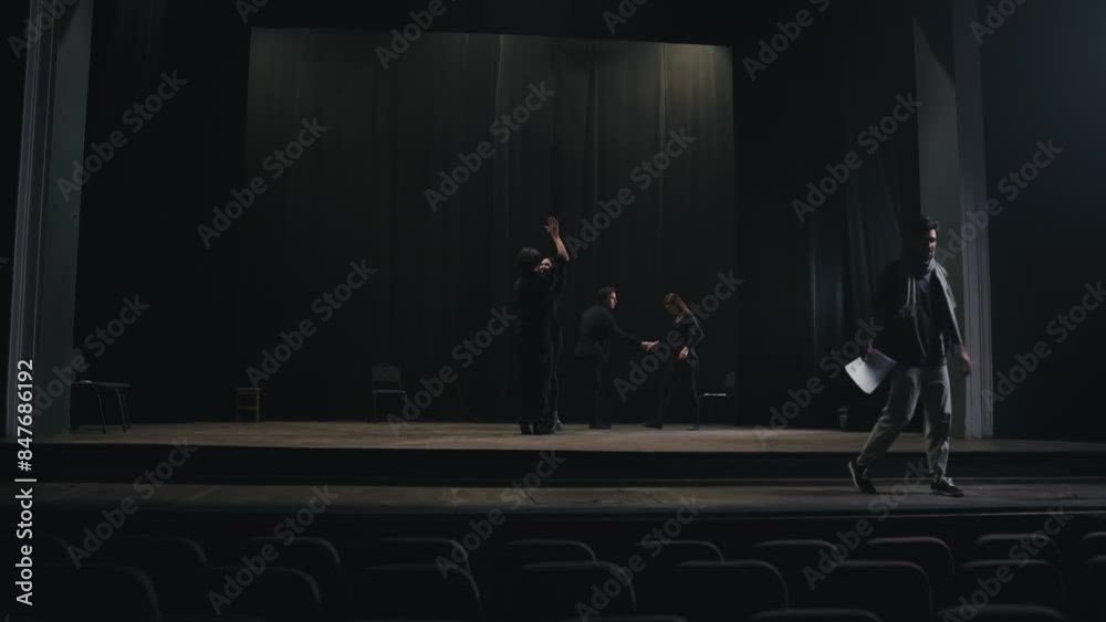 Confident expressive stage director in a gray T-shirt and scarf leaves the stage while his actors in black suits rehearse their roles and script on stage in the theater