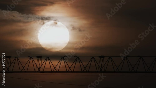 A radiant sun descends behind a metal construction, with faint colored clouds and power lines visible. photo