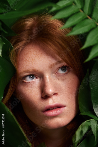 attractive woman with freckles on face and red hair posing with palm leaves, isolated on green, looking into the distance. 