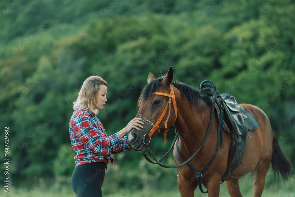Obraz premium Happy blonde with horse in forest. Woman and a horse walking through the field during the day. Dressed in a plaid shirt and black leggings.