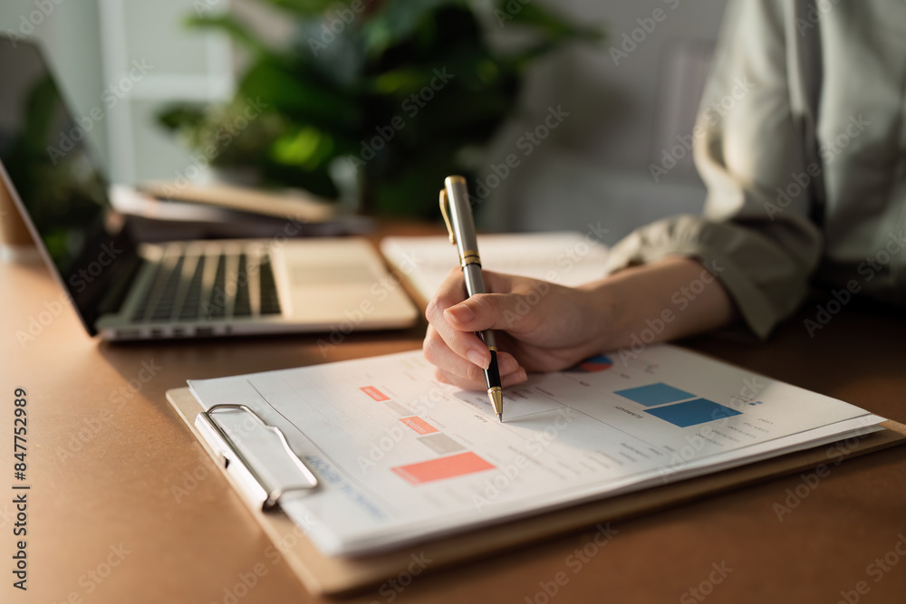 Business Professional Analyzing Financial Data on a Clipboard with Laptop in Background in Modern Office Setting Business Professional Analyzing Financial Data on a Clipboard with Laptop in Background in Modern Office Setting
