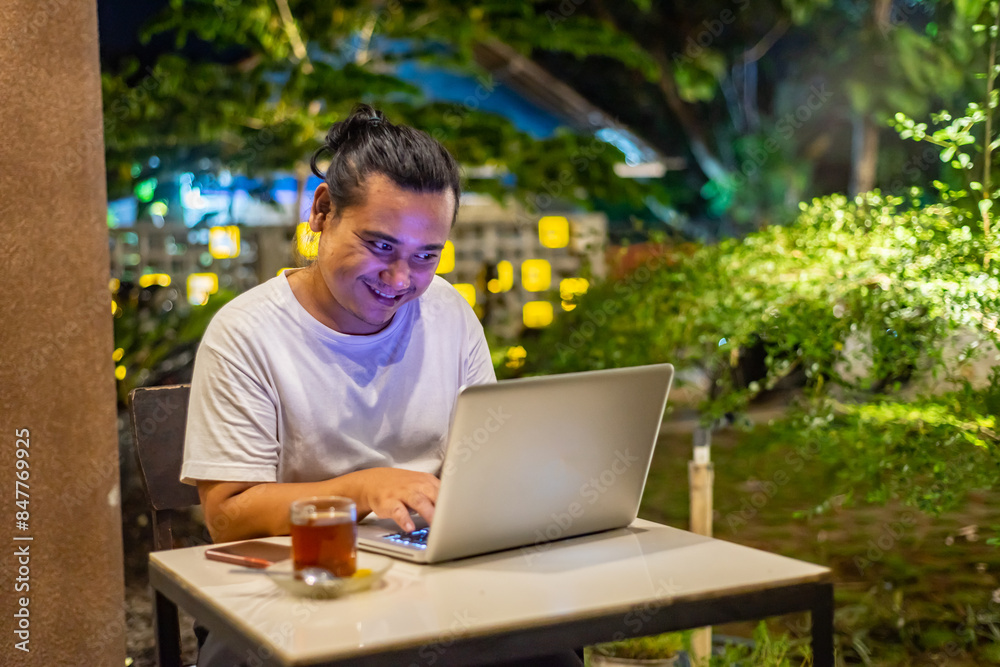 Young Asian man, digital nomad working remotely from cafe, using laptop and smartphone. young Asian men are hanging out in a cafe with a nature theme at night