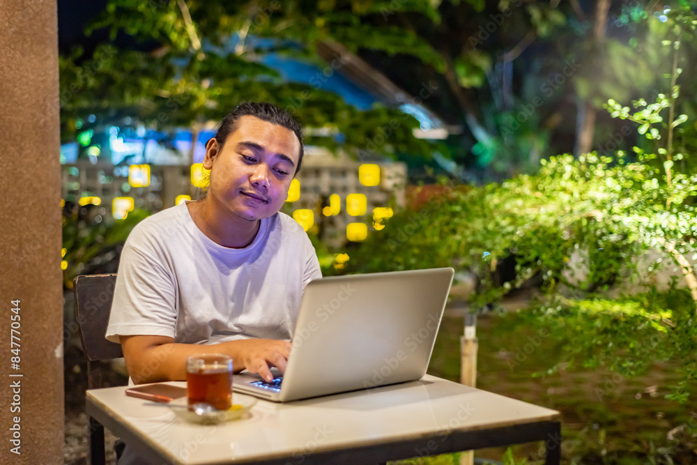 Young Asian man, digital nomad working remotely from cafe, using laptop and smartphone. young Asian men are hanging out in a cafe with a nature theme at night