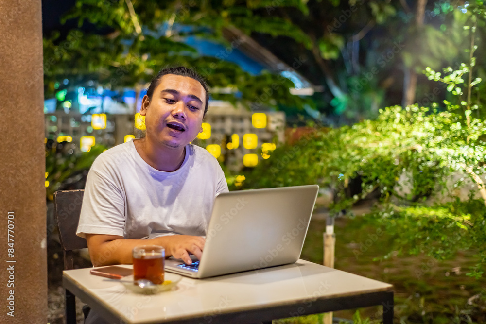 Young Asian man, digital nomad working remotely from cafe, using laptop and smartphone. young Asian men are hanging out in a cafe with a nature theme at night