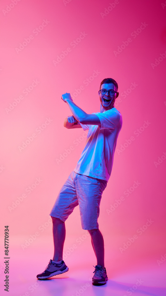 Full-length image of young man in glasses and casual clothes cheerfully dancing and having fun against pink background in neon light. Concept of human emotions, facial expression, lifestyle Full-length image of young man in glasses and casual clothes cheerfully dancing and having fun against pink background in neon light. Concept of human emotions, facial expression, lifestyle