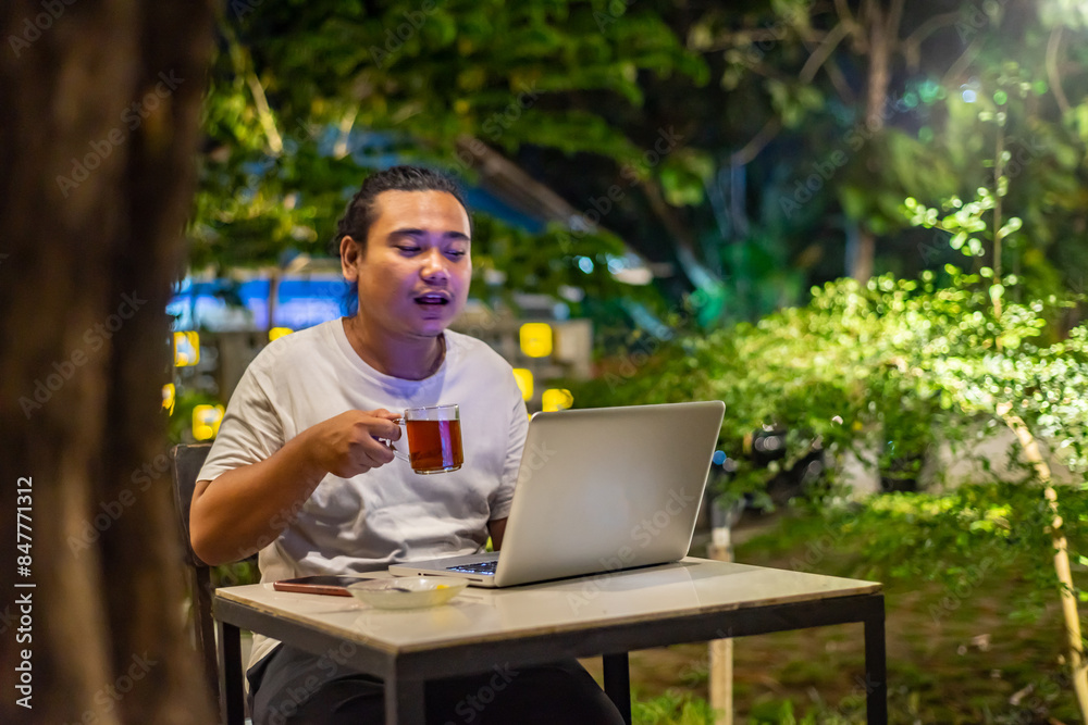 Young Asian man, digital nomad working remotely from cafe, using laptop and smartphone. young Asian men are hanging out in a cafe with a nature theme at night