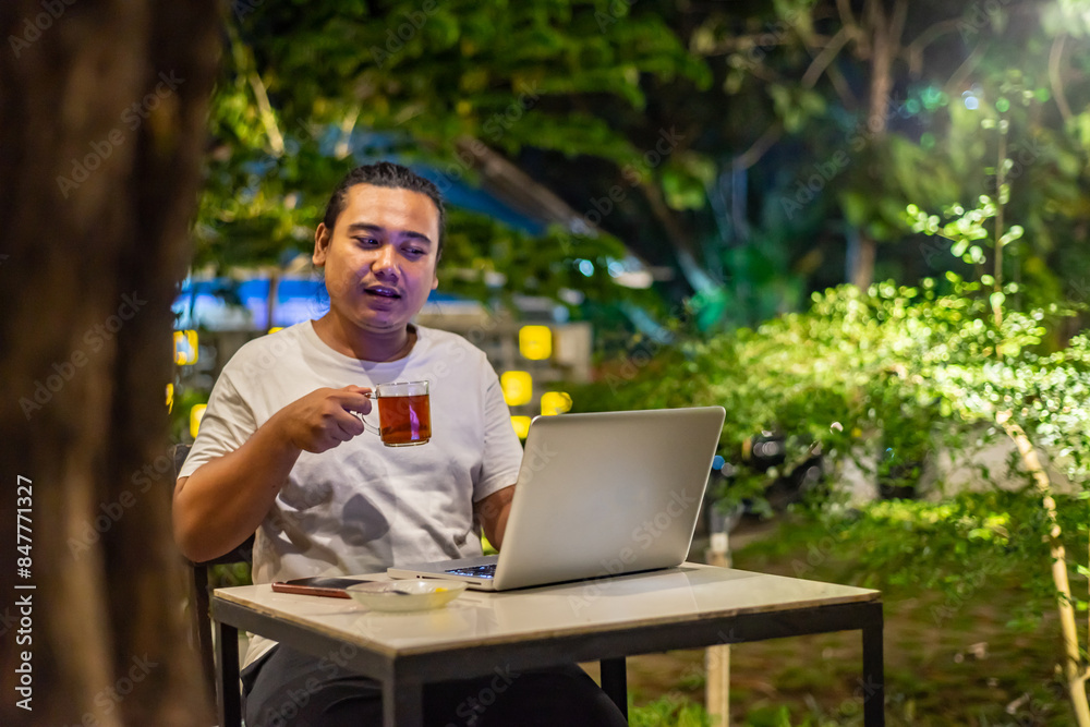 Young Asian man, digital nomad working remotely from cafe, using laptop and smartphone. young Asian men are hanging out in a cafe with a nature theme at night