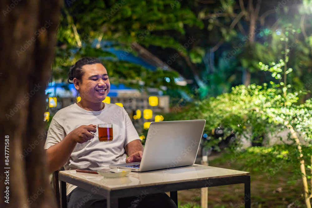 Young Asian man, digital nomad working remotely from cafe, using laptop and smartphone. young Asian men are hanging out in a cafe with a nature theme at night