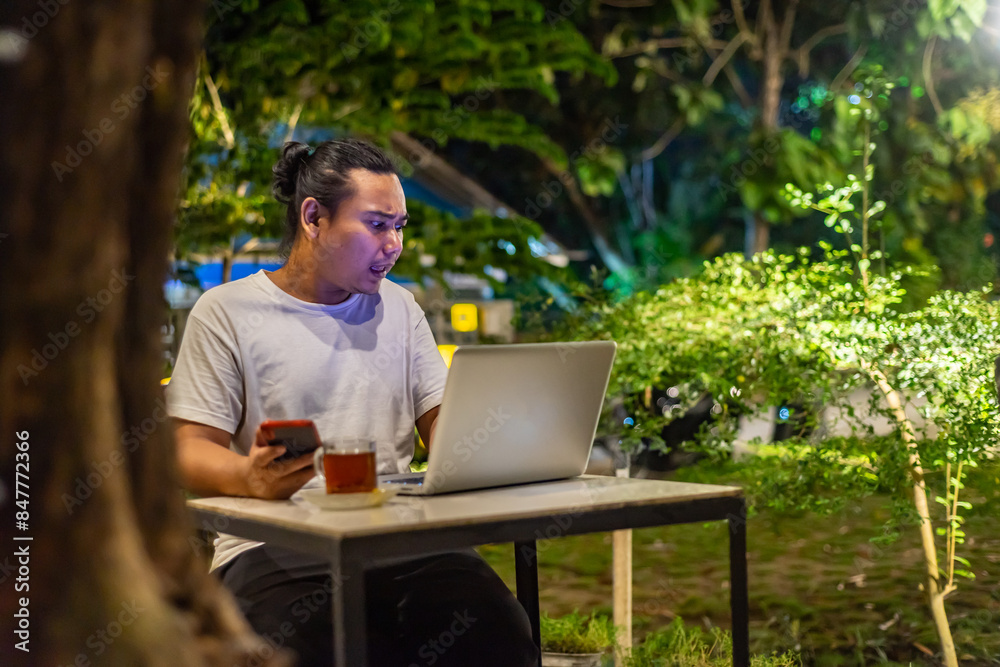 Young Asian man, digital nomad working remotely from cafe, using laptop and smartphone. young Asian men are hanging out in a cafe with a nature theme at night