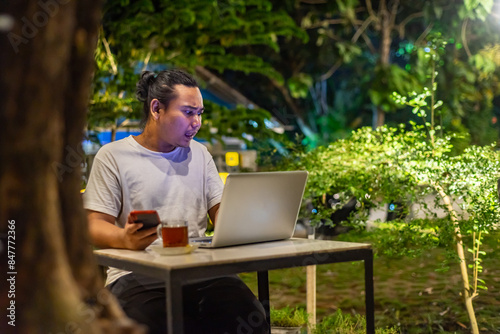 Obraz na płótnie Young Asian man, digital nomad working remotely from cafe, using laptop and smartphone. young Asian men are hanging out in a cafe with a nature theme at night w salonie