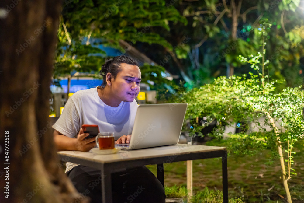 Young Asian man, digital nomad working remotely from cafe, using laptop and smartphone. young Asian men are hanging out in a cafe with a nature theme at night