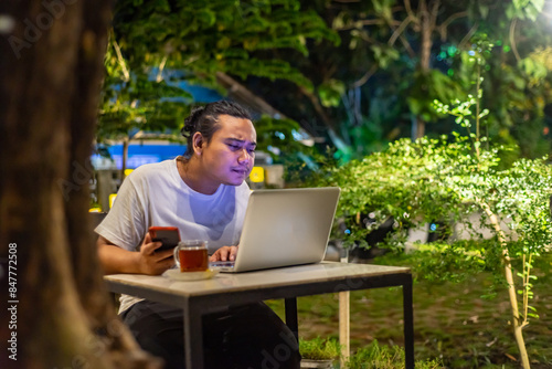 Obraz na płótnie Young Asian man, digital nomad working remotely from cafe, using laptop and smartphone. young Asian men are hanging out in a cafe with a nature theme at night w salonie