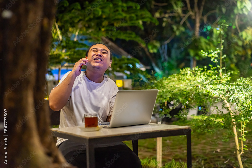 Young Asian man, digital nomad working remotely from cafe, using laptop and smartphone. young Asian men are hanging out in a cafe with a nature theme at night
