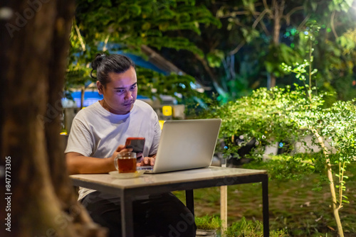 Obraz na płótnie Young Asian man, digital nomad working remotely from cafe, using laptop and smartphone. young Asian men are hanging out in a cafe with a nature theme at night w salonie