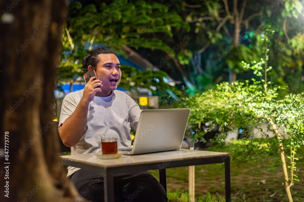 Young Asian man, digital nomad working remotely from cafe, using laptop and smartphone. young Asian men are hanging out in a cafe with a nature theme at night