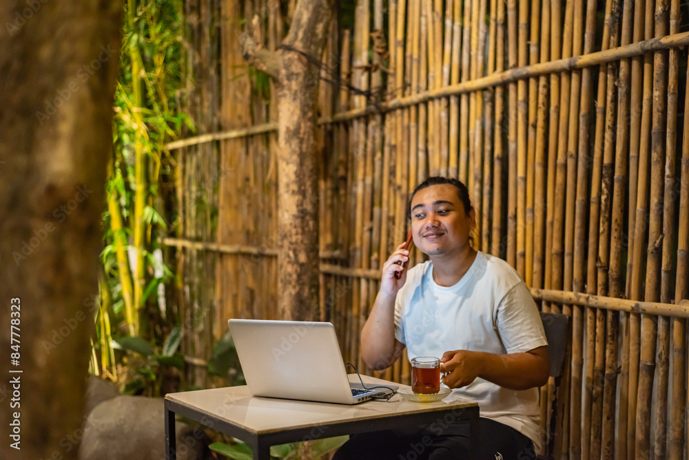 Young Asian man, digital nomad working remotely from cafe, using laptop and smartphone. young Asian men are hanging out in a cafe with a nature theme at night