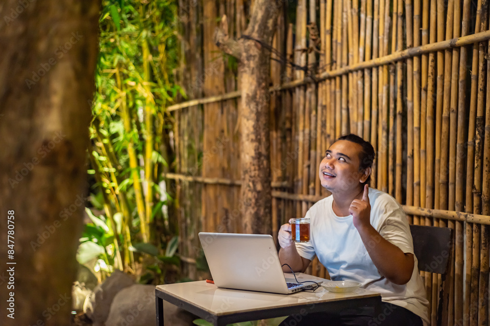 Young Asian man, digital nomad working remotely from cafe, using laptop and smartphone. young Asian men are hanging out in a cafe with a nature theme at night