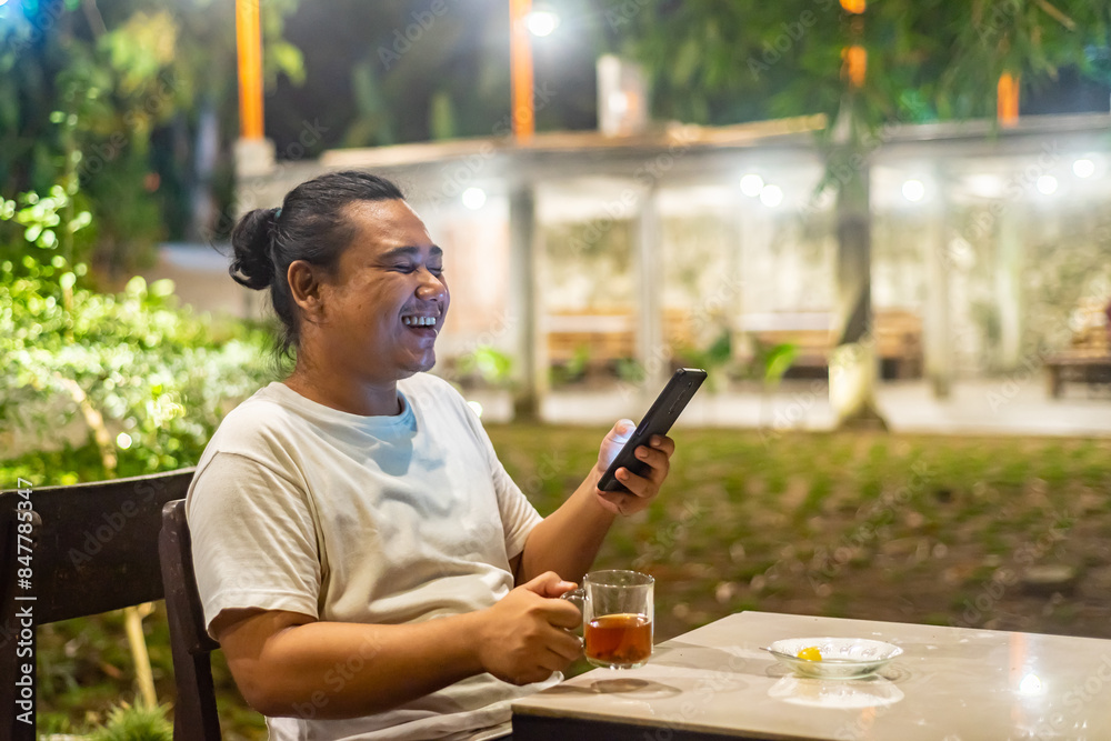 Young Asian man, digital nomad working remotely from cafe, using laptop and smartphone. young Asian men are hanging out in a cafe with a nature theme at night