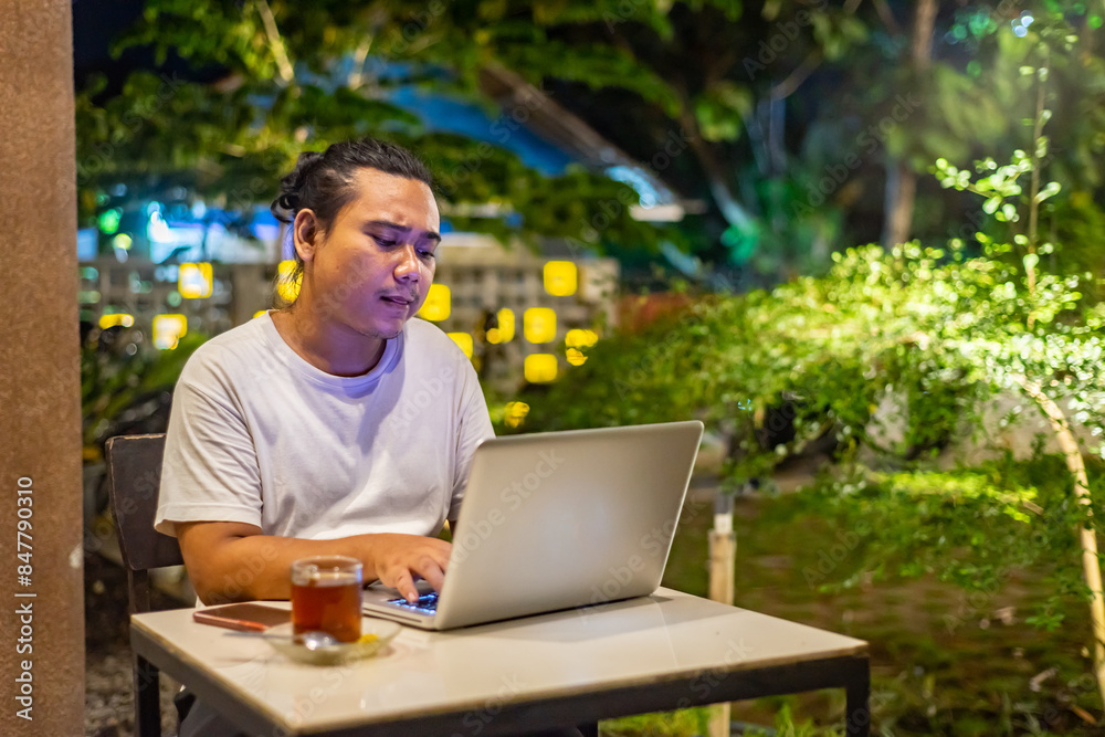Young Asian man, digital nomad working remotely from cafe, using laptop and smartphone. young Asian men are hanging out in a cafe with a nature theme at night