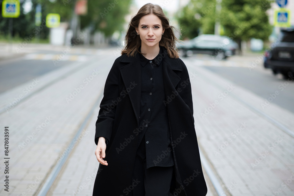 Fototapeta premium Attractive caucasian young woman in black coat walking down city street on warm autumn day. Outdoors portrait. Model in stylish clothes. Lifestyle, fashion concept.