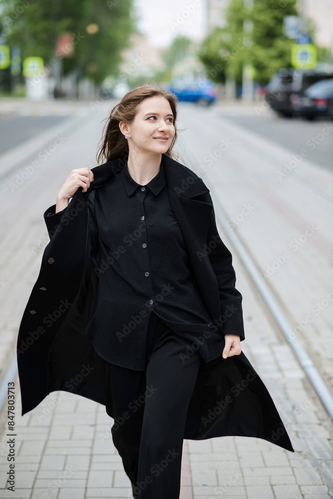 Attractive caucasian young woman in black coat walking down city street on warm autumn day. Outdoors portrait. Model in stylish clothes. Lifestyle, fashion concept. Attractive caucasian young woman in black coat walking down city street on warm autumn day. Outdoors portrait. Model in stylish clothes. Lifestyle, fashion concept.